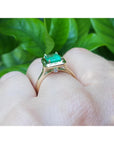 On a hand the underside view of an emerald solitaire ring in yellow gold against a leafy background.