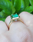 On a hand the underside view of an emerald solitaire ring in yellow gold against a leafy background.