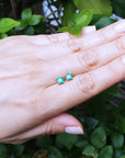 Hand displaying a pair of 14k gold emerald stud earrings against a leafy background.