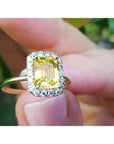 Gold ring with a yellow sapphire gemstone held between fingers against a blurred natural background