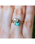 A gold ring with a rectangular clear gemstone and an emerald-cut turquoise green gemstone, shown worn on a person's finger.