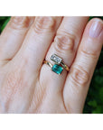 A close-up of a hand wearing a gold ring set with a rectangular clear gemstone and a larger rectangular emerald-cut green gemstone, against a blurred green background.