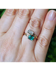A close-up of a hand wearing a 18k yellow gold toi et moi emerald and diamond ring, against a blurred green background.