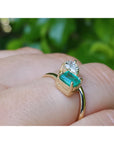 A yellow gold ring with a rectangular emerald-cut emerald and a pear cut diamond, shown worn on a person's finger against a blurred green background.