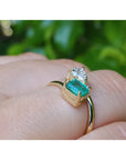 A yellow gold ring with a rectangular emerald-cut emerald and a pear cut diamond, shown worn on a person's finger against a blurred green background.