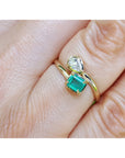 A close-up of a gold you and me ring with a square emerald-cut emerald and a smaller diamond, worn on a finger.