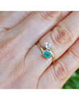 A gold ring with a square-cut emerald gemstone and a small pear cut diamond, shown worn on a person's finger.