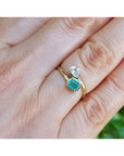 A gold ring with a square-cut emerald gemstone and a small pear cut diamond, shown worn on a person's finger.