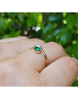 A gold ring with a pear-cut natural emerald, worn on a person's finger against a blurred background of green foliage.