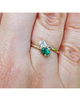 A gold ring with a diamond pear-cut gemstone and a natural emerald gemstone, shown worn on a person's finger.