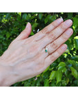 A You and Me 18k gold ring with a pear cut emerald and and diamond worn on the ring finger of a hand against a background of green foliage.