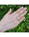 A You and Me 18k gold ring with a pear cut emerald and and diamond worn on the ring finger of a hand against a background of green foliage.