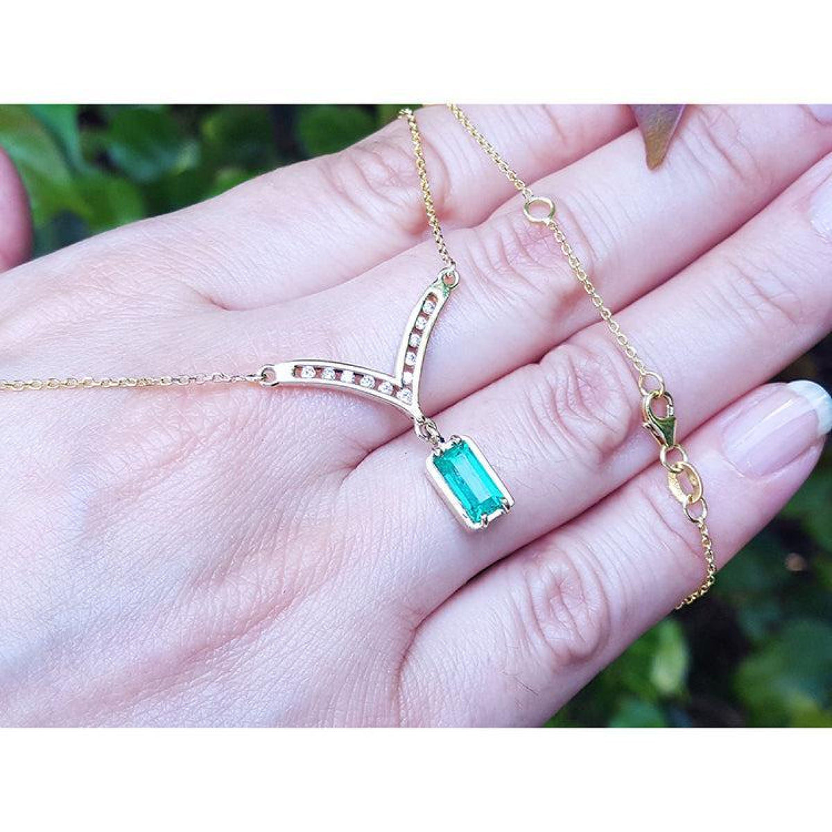 14K yellow gold necklace with a baguette-cut Colombian emerald dangling from a diamond-studded curved V, shown resting on a person's hand against a blurred green background.