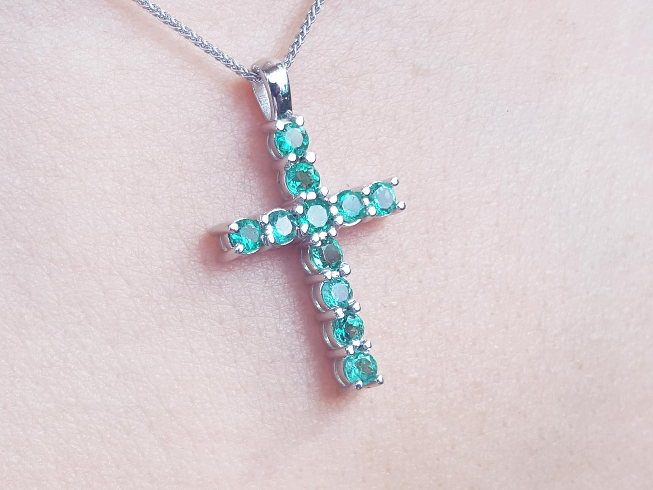 White gold emerald cross necklace on a light pink skin background.