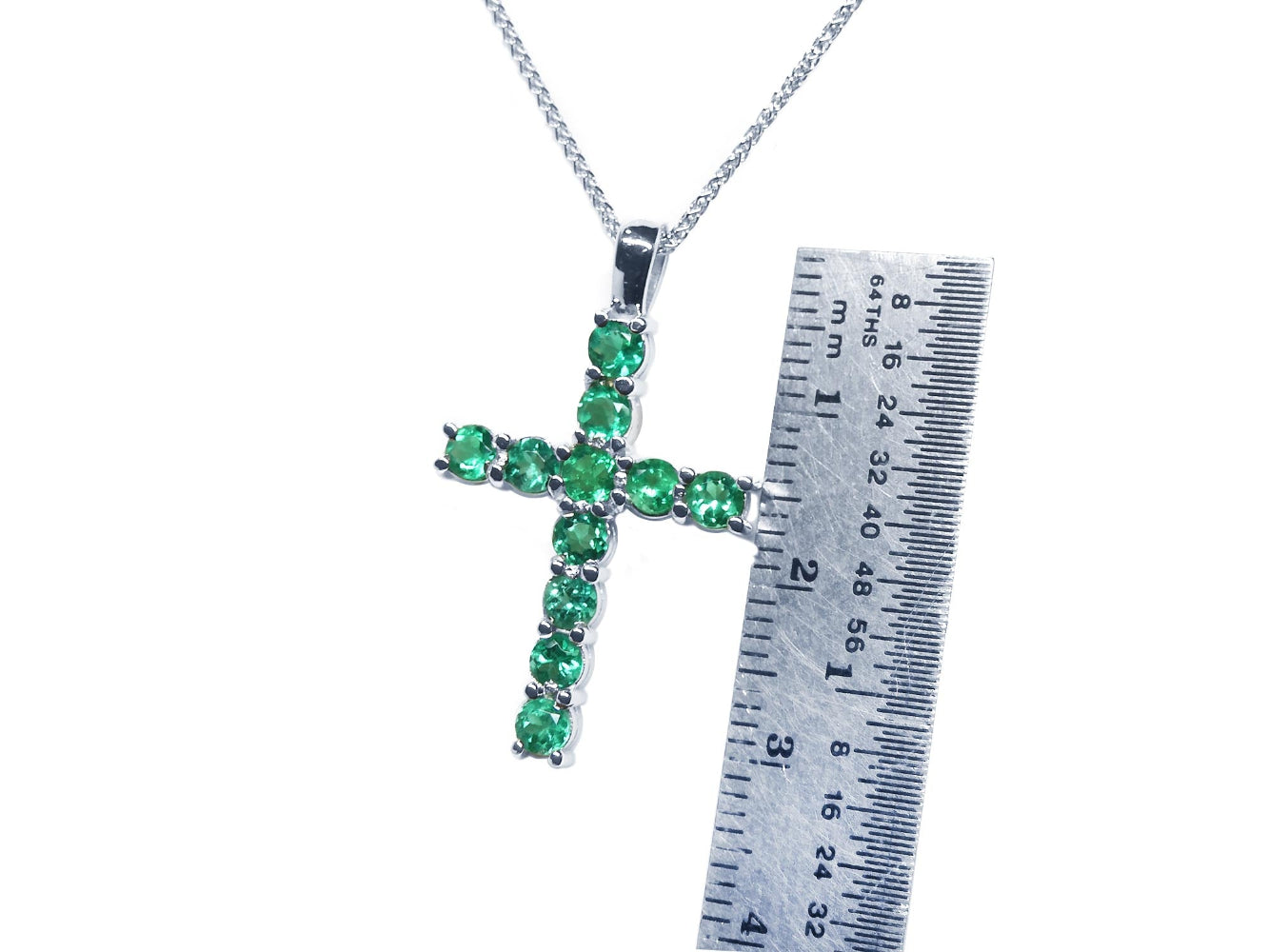 A natural emerald cross pendant on a white gold chain with a ruler for scale.