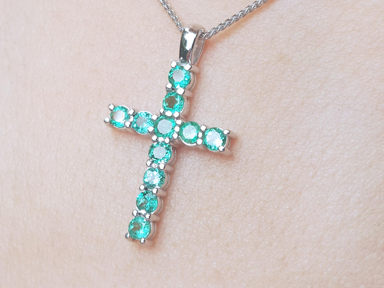 Cross-shaped Muzo emeralds cross pendant on a chain against a light skin background.