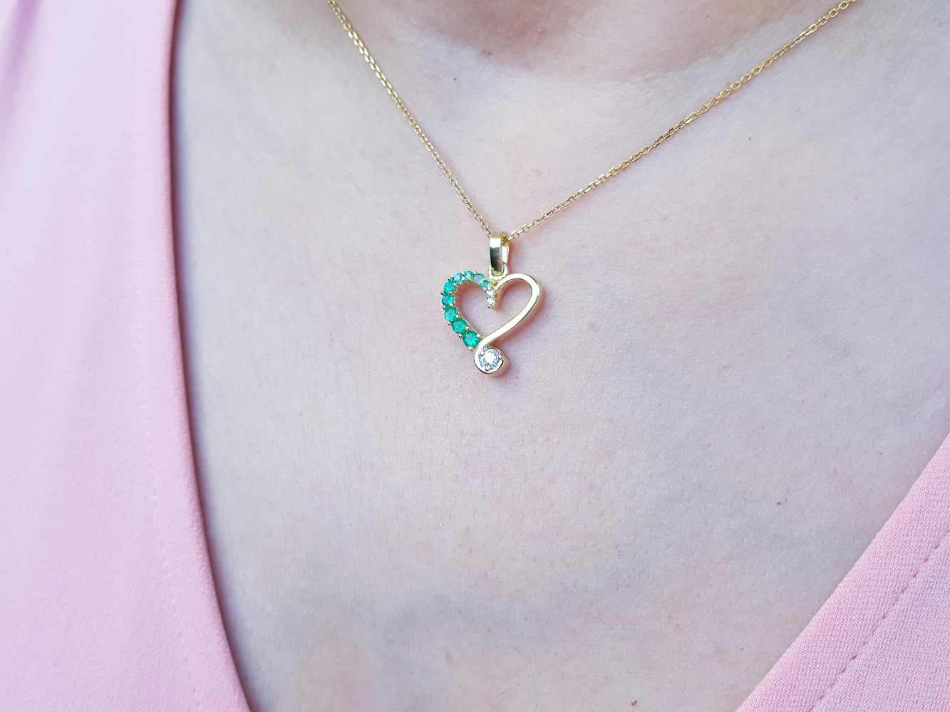Heart-shaped pendant with natural emeralds on a gold chain, worn by a person against a pink background.
