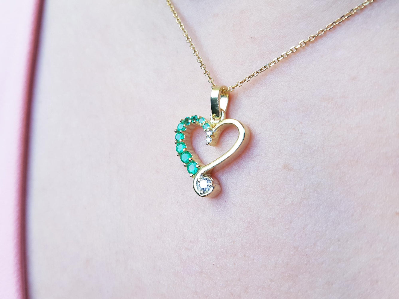 Heart-shaped pendant with emeralds and a diamond on a gold chain against a pink background