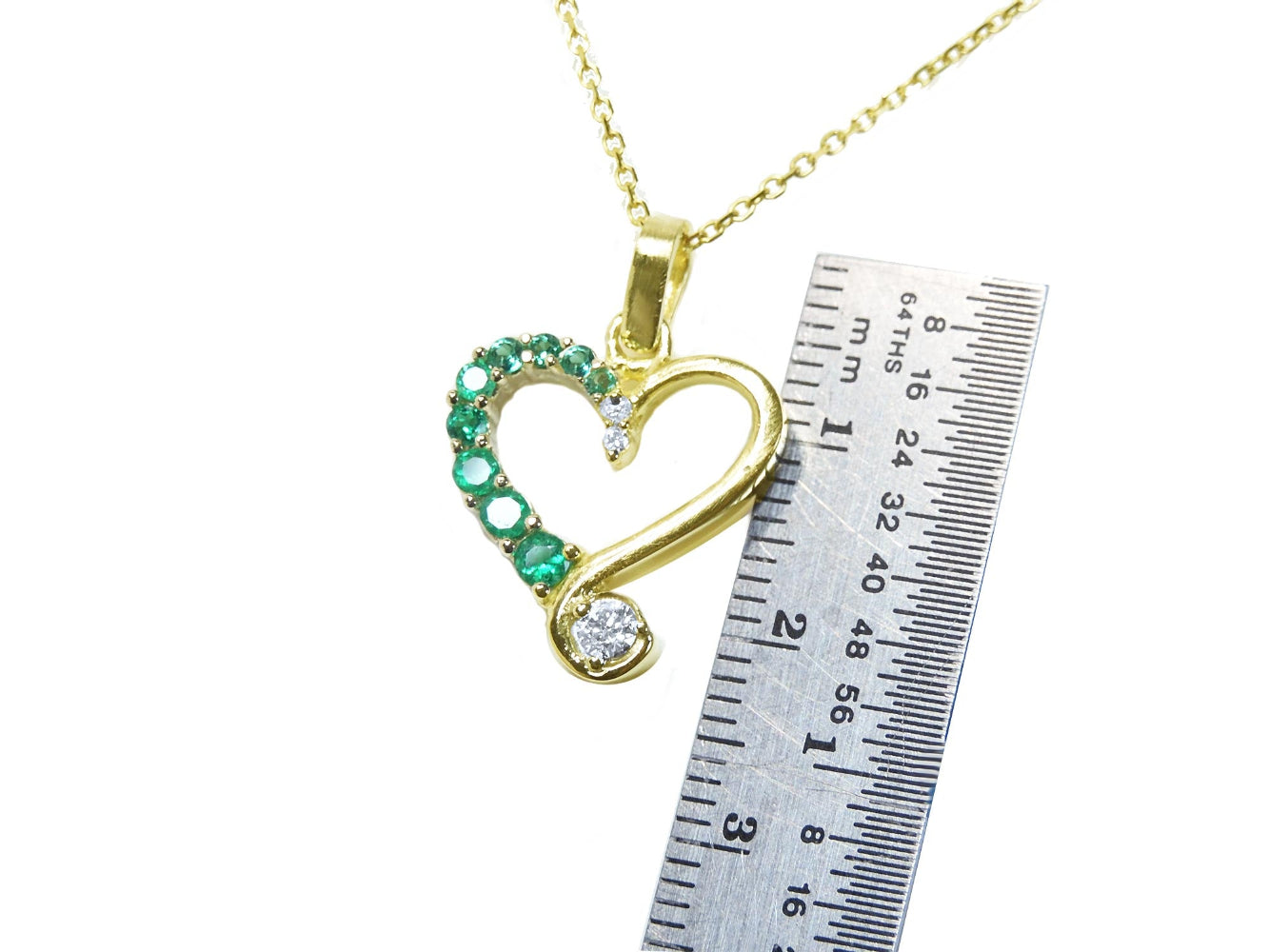 Gold heart-shaped pendant with emeralds and diamonds on a gold chain, next to a ruler for scale.