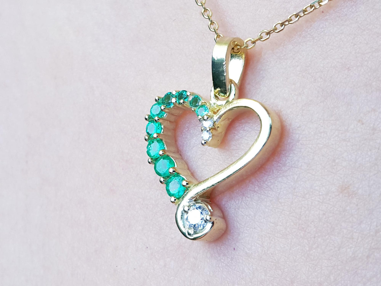 Heart-shaped gold pendant with emerald gemstones on a light pink background.