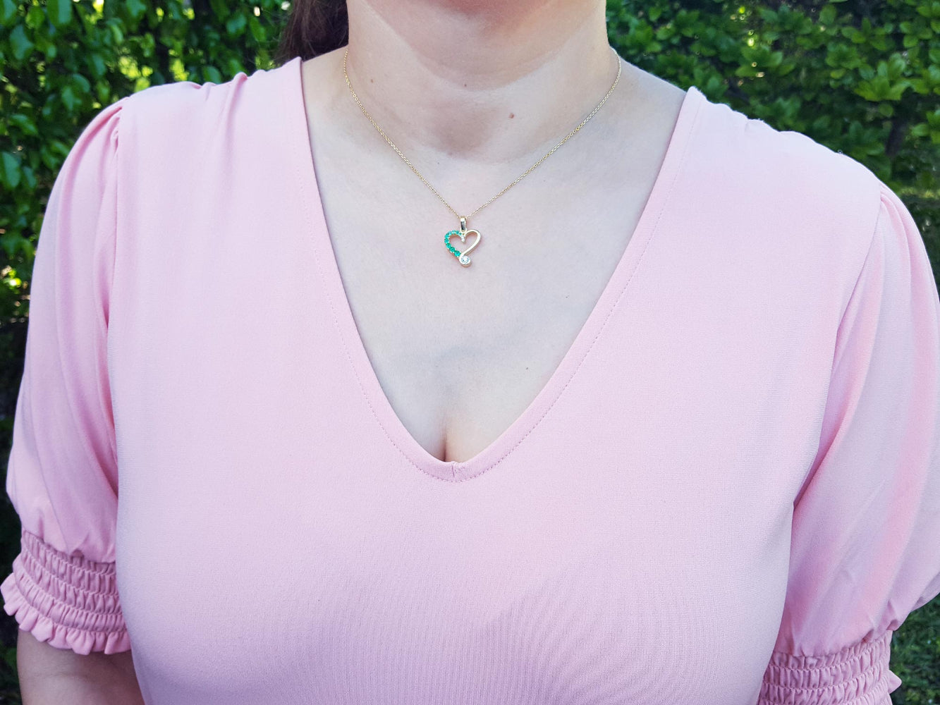 A woman wearing a light pink blouse and a heart emerald pendant with a blurred green background.