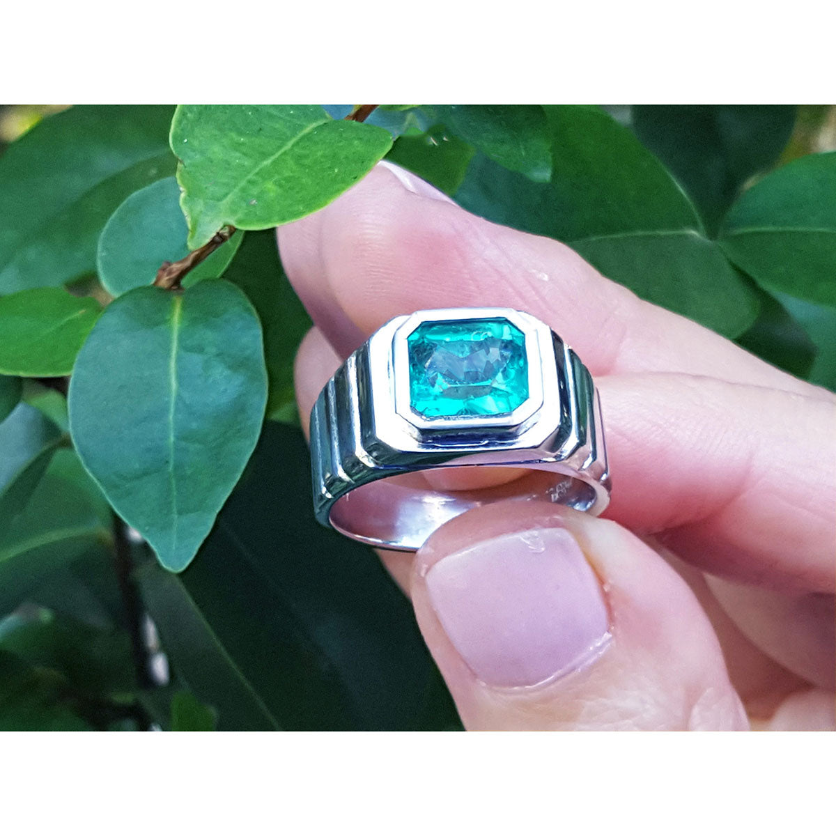 A bezel set natural colombian emerald solitaire ring for men held by a hand against a green leafy background.