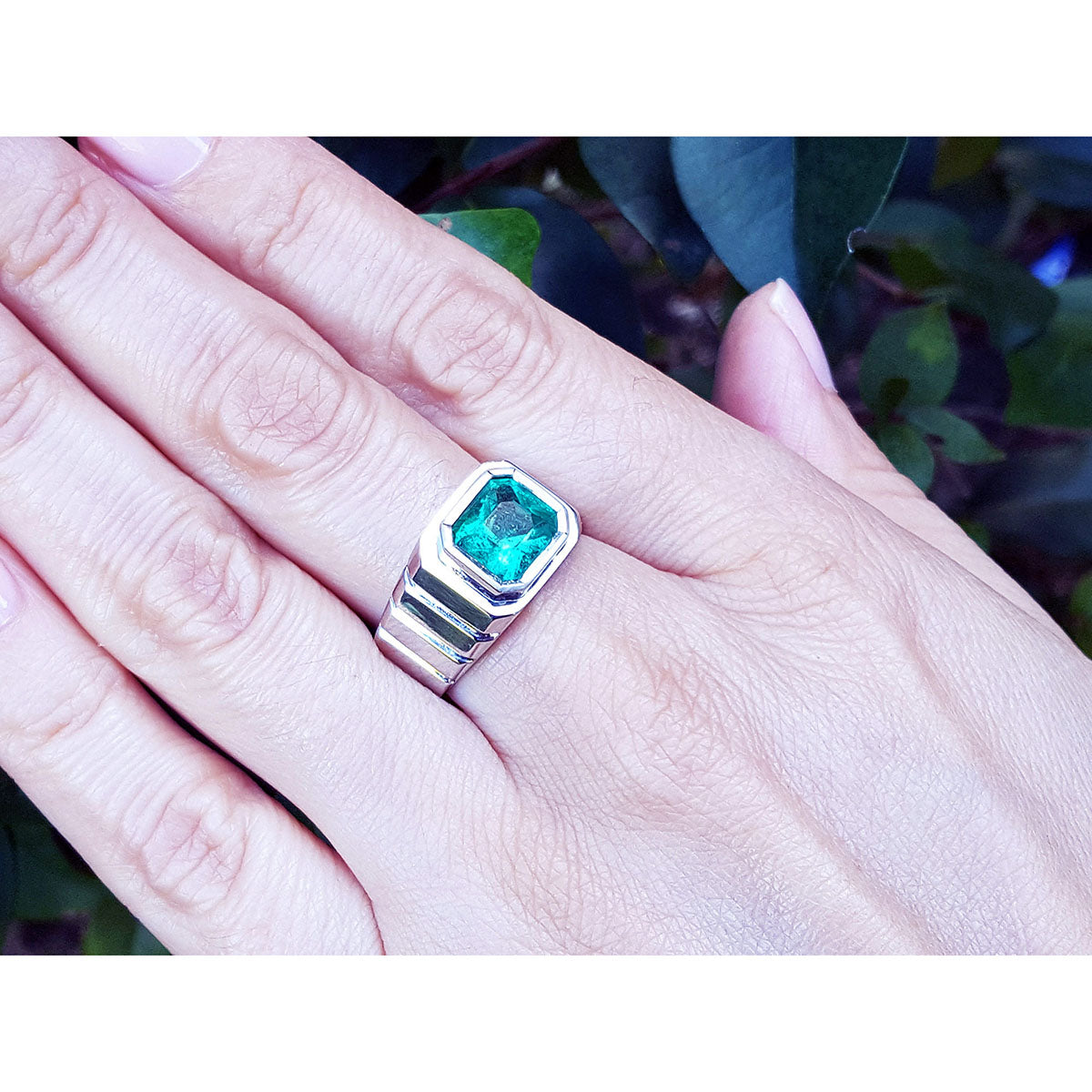 A men&#39;s white gold solitaire emerald ring on a hand against a blurred natural background.