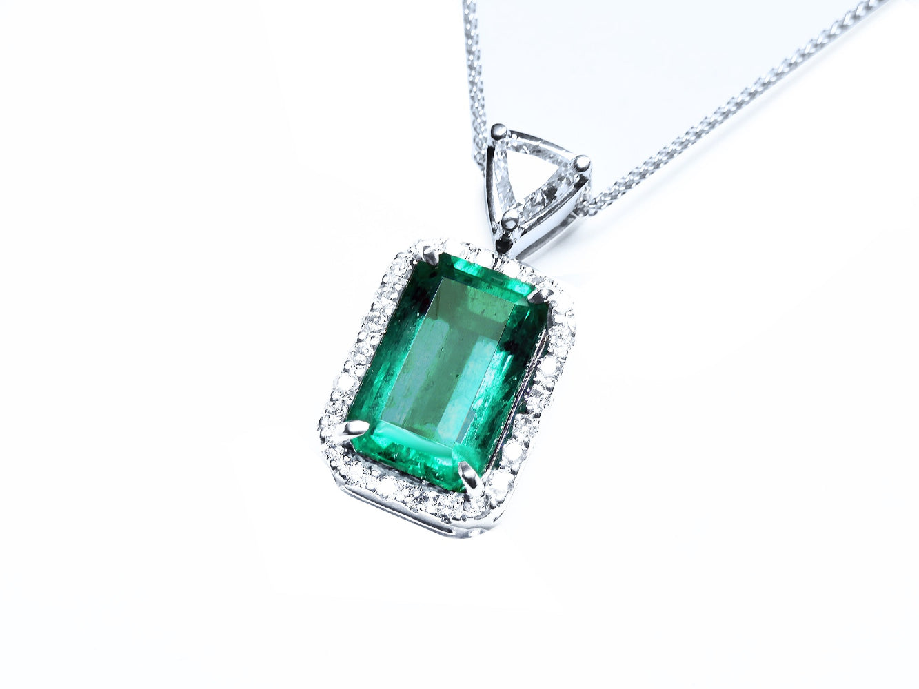 A rectangular emerald encircled by a halo diamond pendant with a top bail with trillion-cut diamond in white gold.
