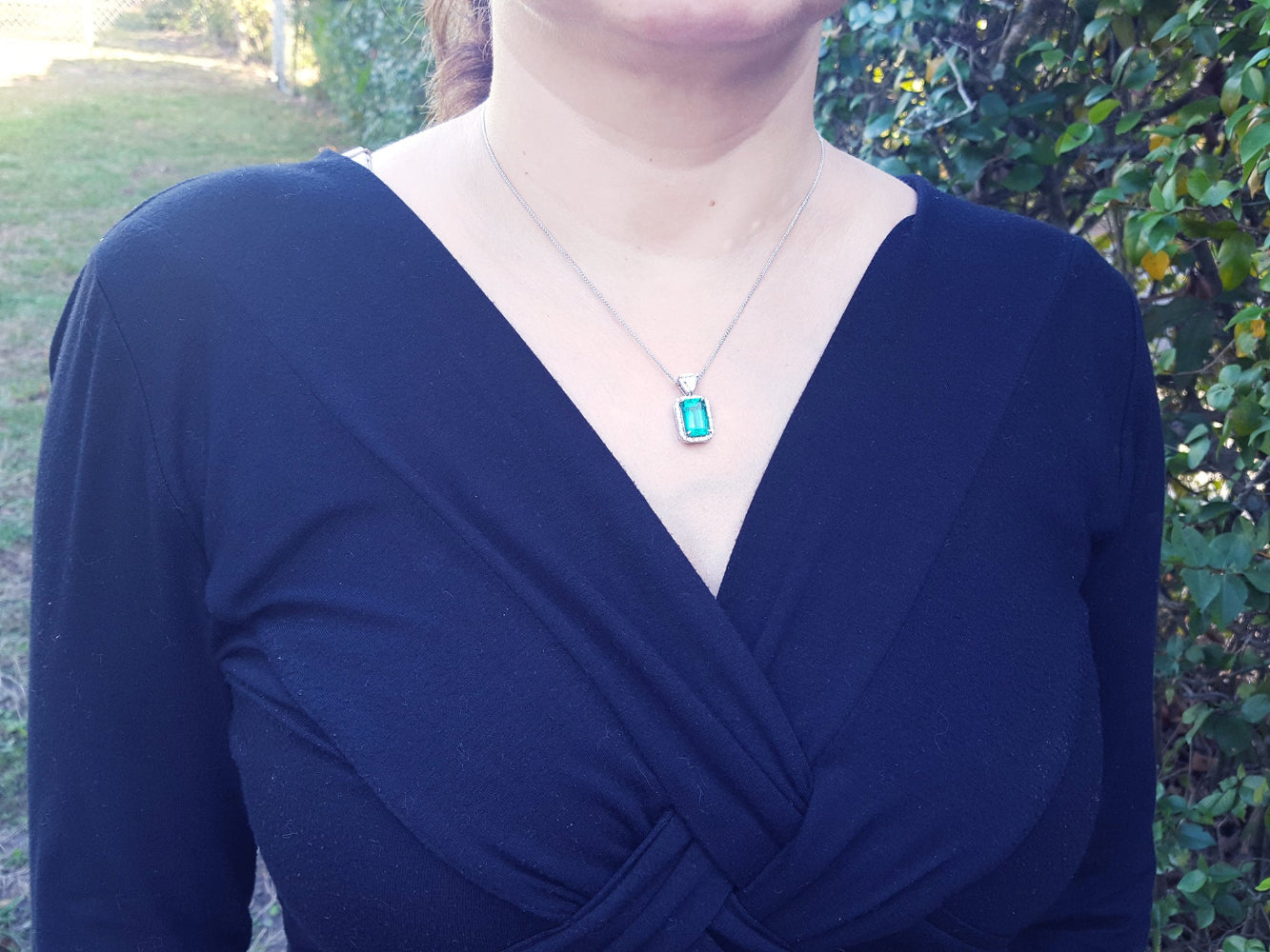 A woman wearing a blue dress with a necklace featuring a natural emerald pendant outdoors.