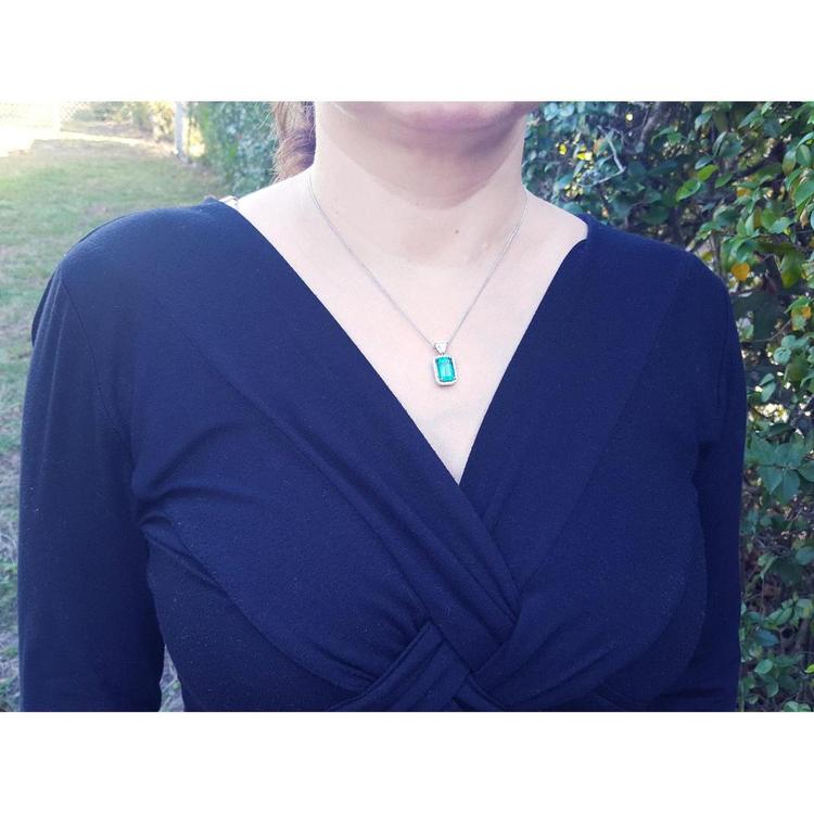 A woman wearing a blue dress with a necklace featuring a natural emerald pendant outdoors.