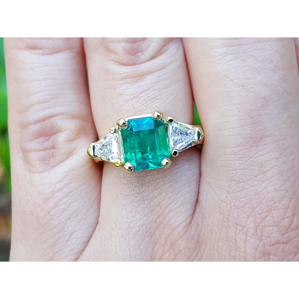 A Gia certified emerald ring in 18k yellow gold and trillion cut side diamond accents on a finger.
