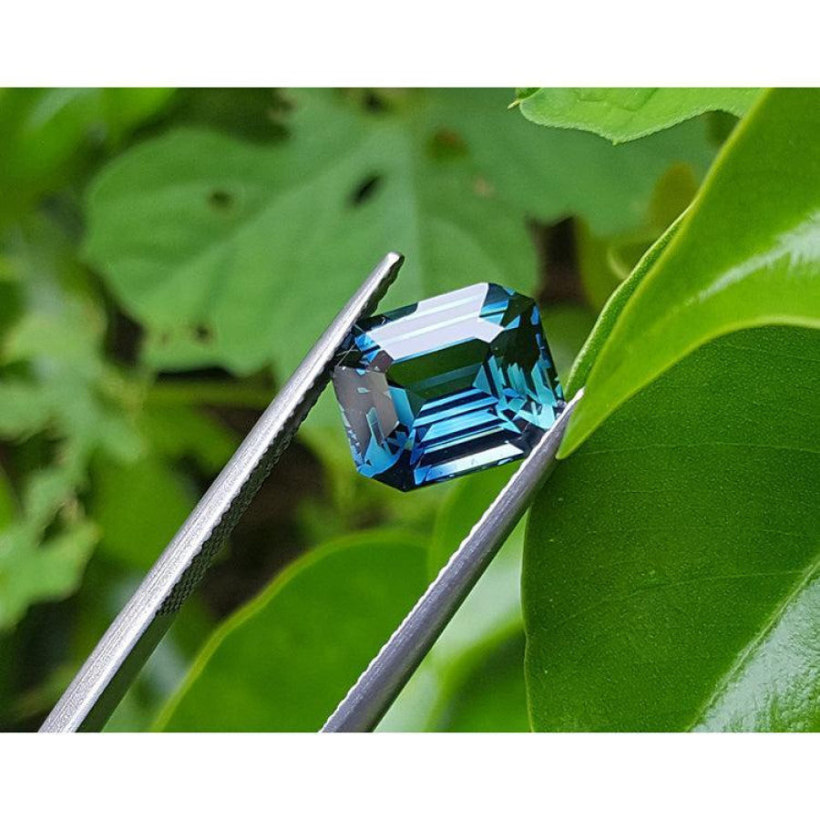GIA certified 6.07 ct. rectangular emerald-cut blue-green sapphire held by tweezers against green leaves.