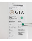 A GIA Emerald Origin Report card for a loose green emerald stone, with a small green gemstone resting on the right side of the white paper.