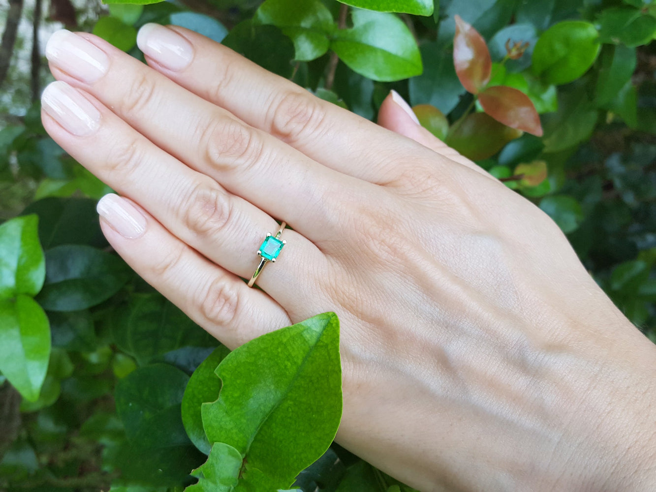 Hand wearing a minimalist emerald solitaire ring representing May&#39;s birthstone jewelry with a green leafy background.