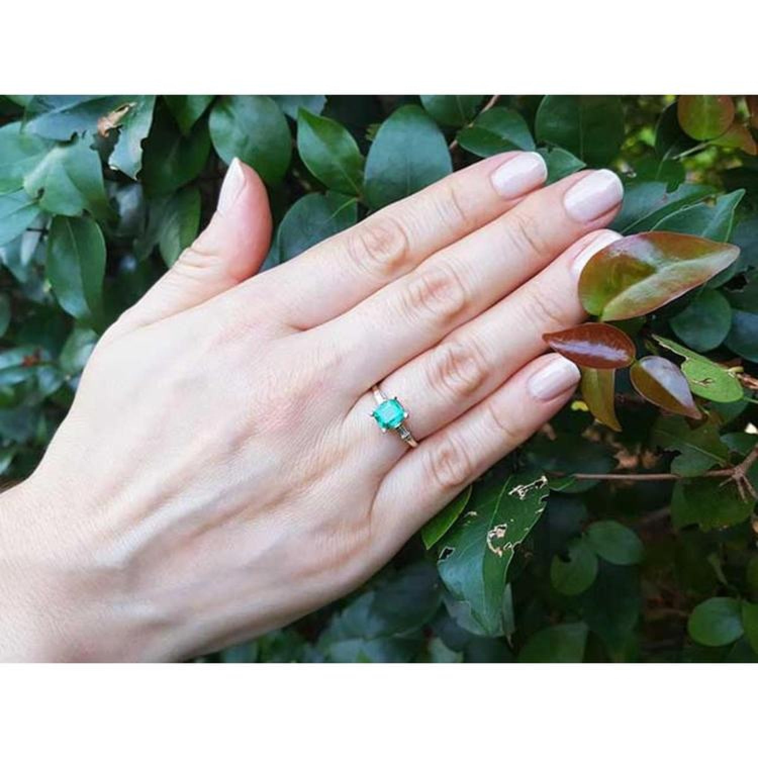 14K yellow gold emerald ring with baguette side diamonds on a hand, against green leaves.