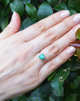 Hand wearing a yellow gold emerald ring against a leafy background.