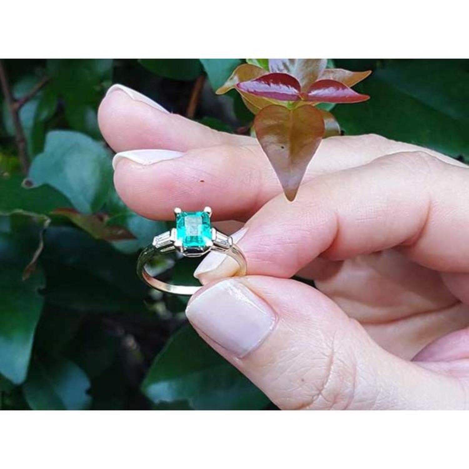 14K yellow gold emerald ring with baguette side diamonds, held against green foliage.