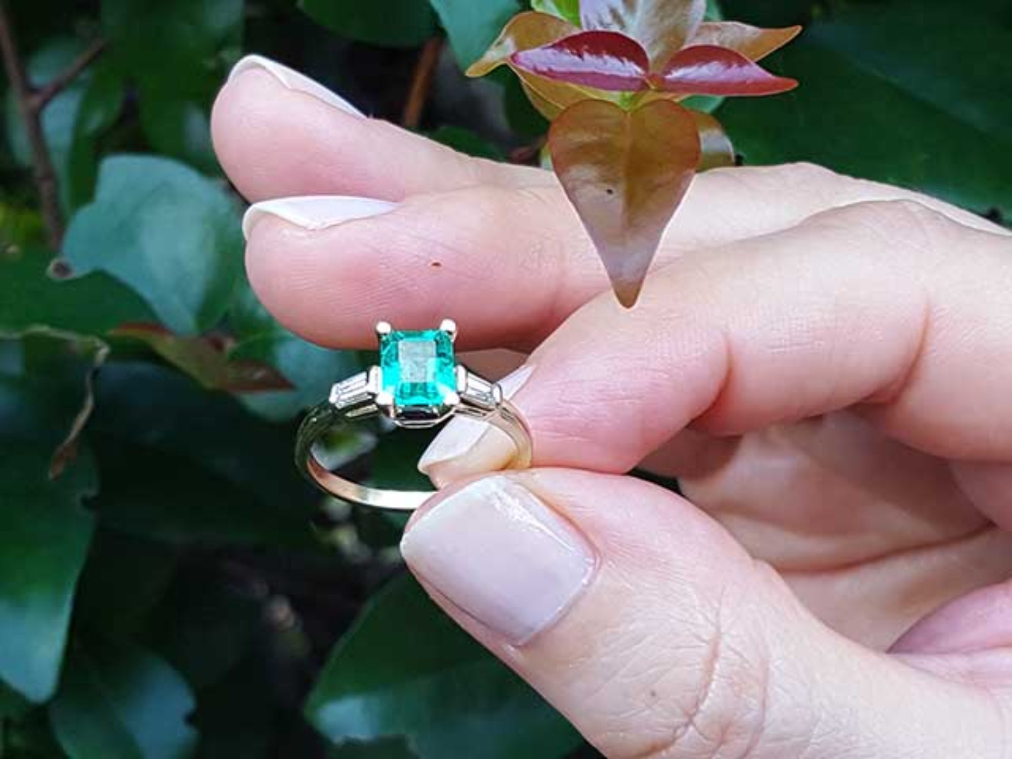 Hand holding a ring with a natural colombian emerald against a leafy background.