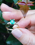 Hand holding a ring with a natural colombian emerald against a leafy background.