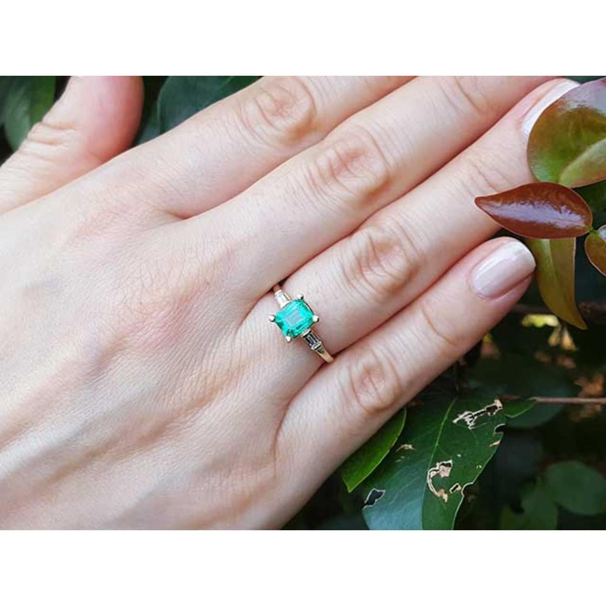 Hand wearing a, emerald May's birthstone ring against a leafy background.