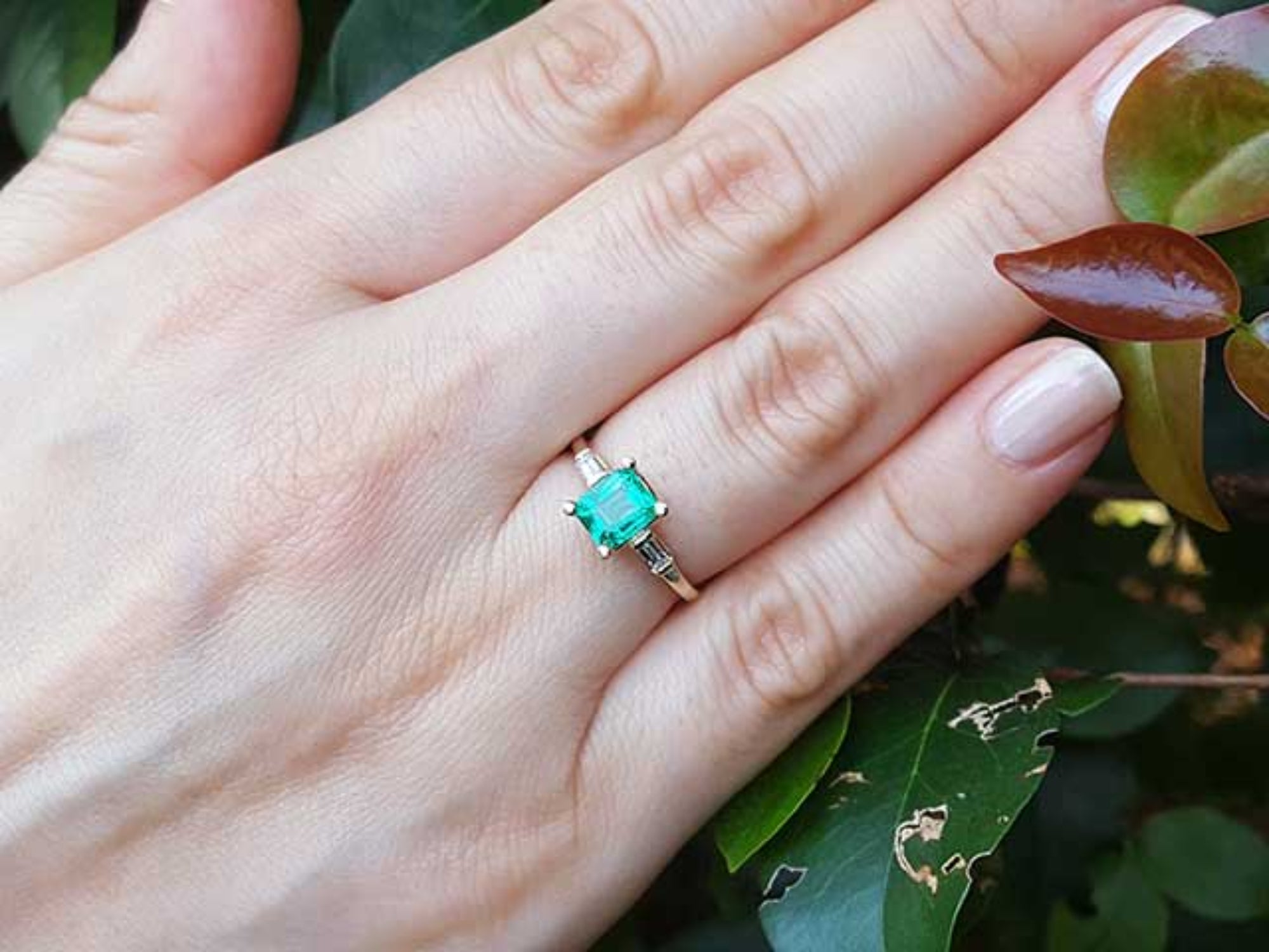 Hand wearing a, emerald May&#39;s birthstone ring against a leafy background.