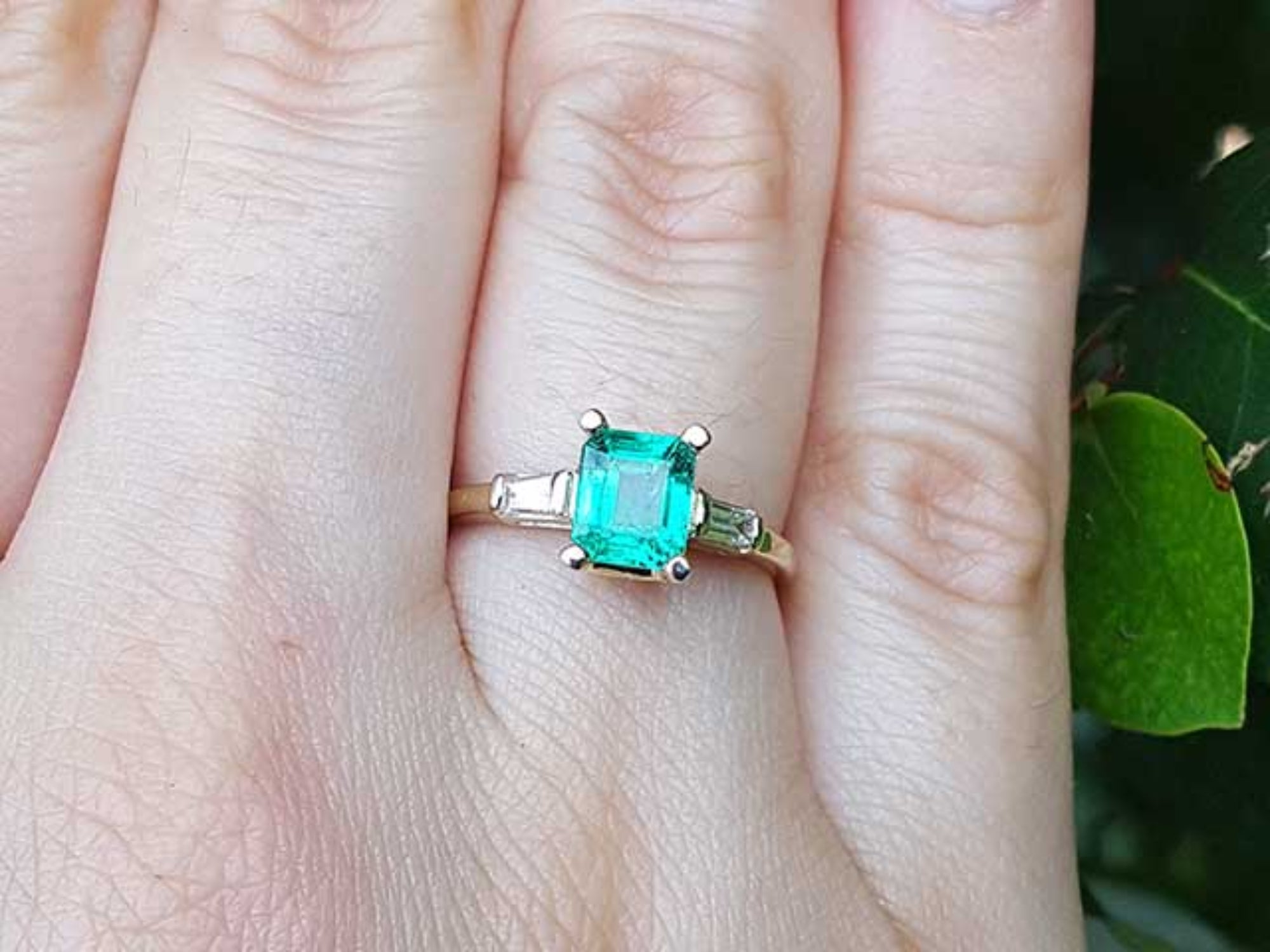 A yellow gold emerald ring with side diamonds on a person's finger, with a natural background.