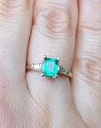 A yellow gold emerald ring with side diamonds on a person's finger, with a natural background.