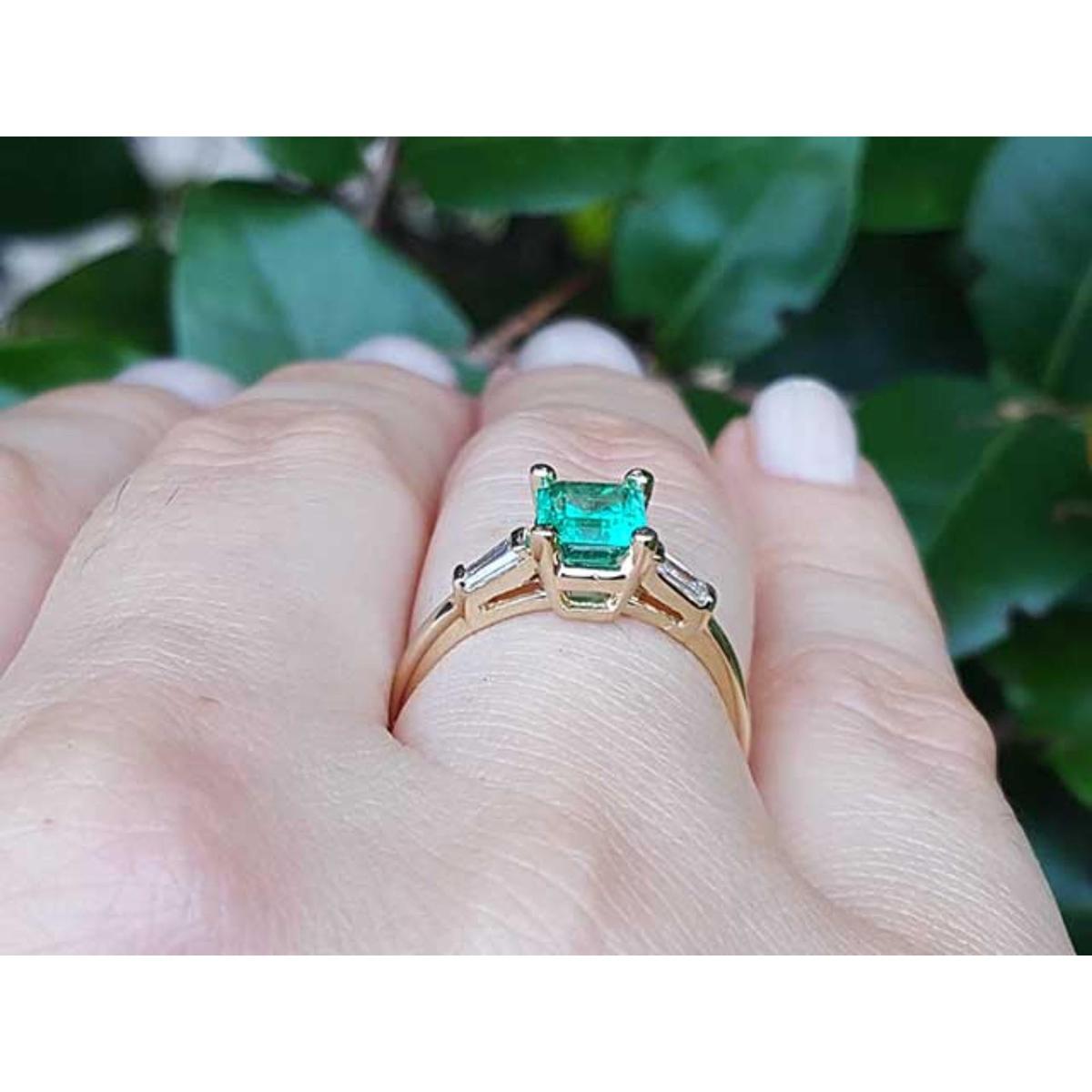 Gold ring with a natural emerald on a hand against a leafy background.