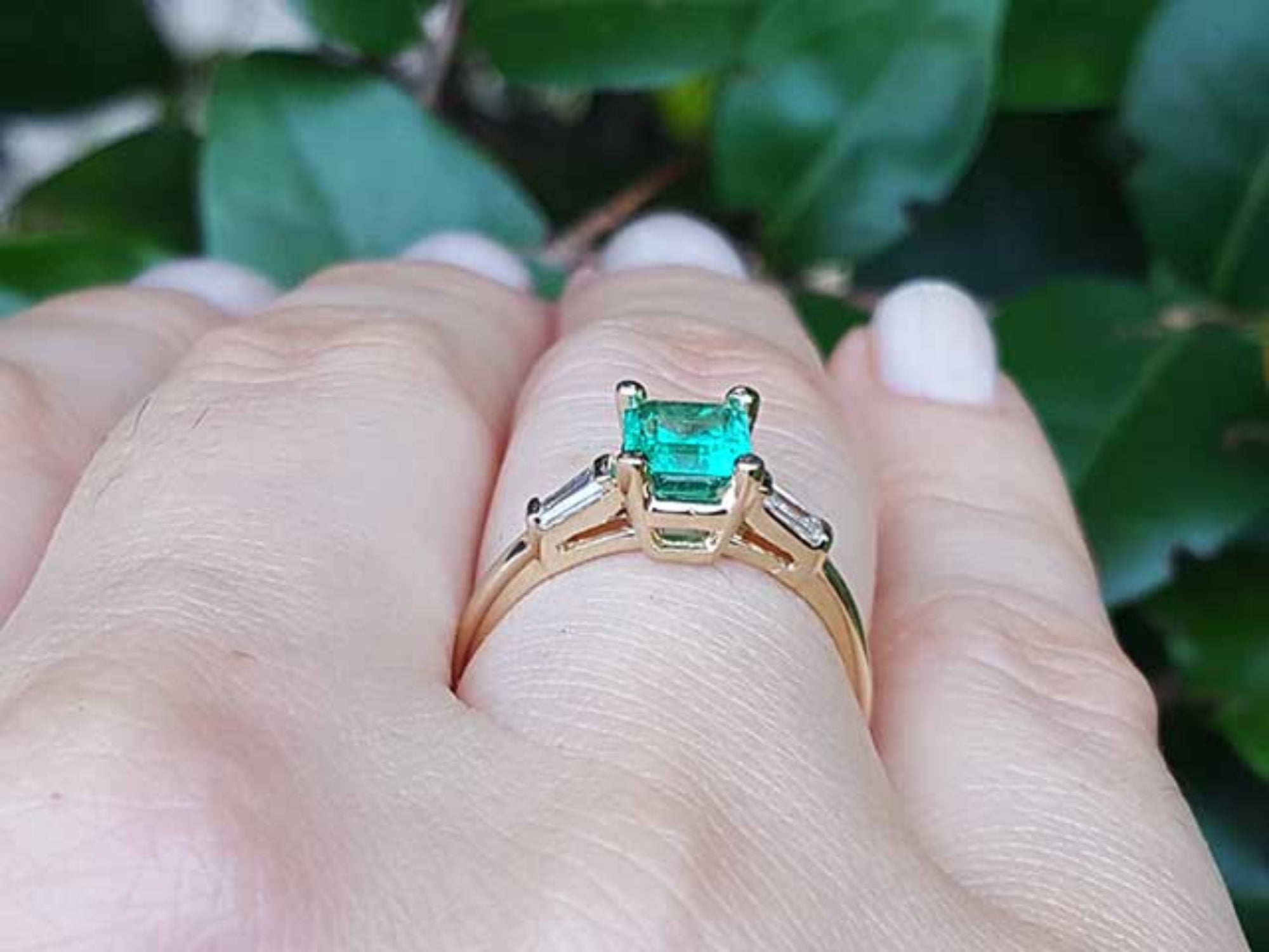 Gold ring with a natural emerald on a hand against a leafy background.