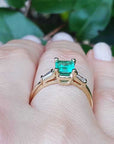Gold ring with a natural emerald on a hand against a leafy background.