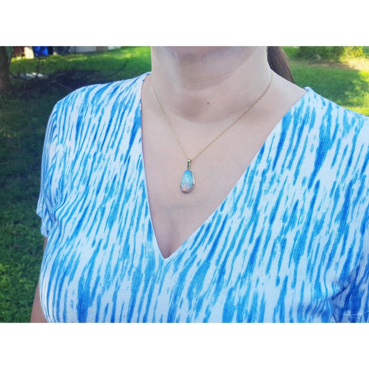A woman wearing a blue and white patterned top with a gold necklace featuring an opal pendant.
