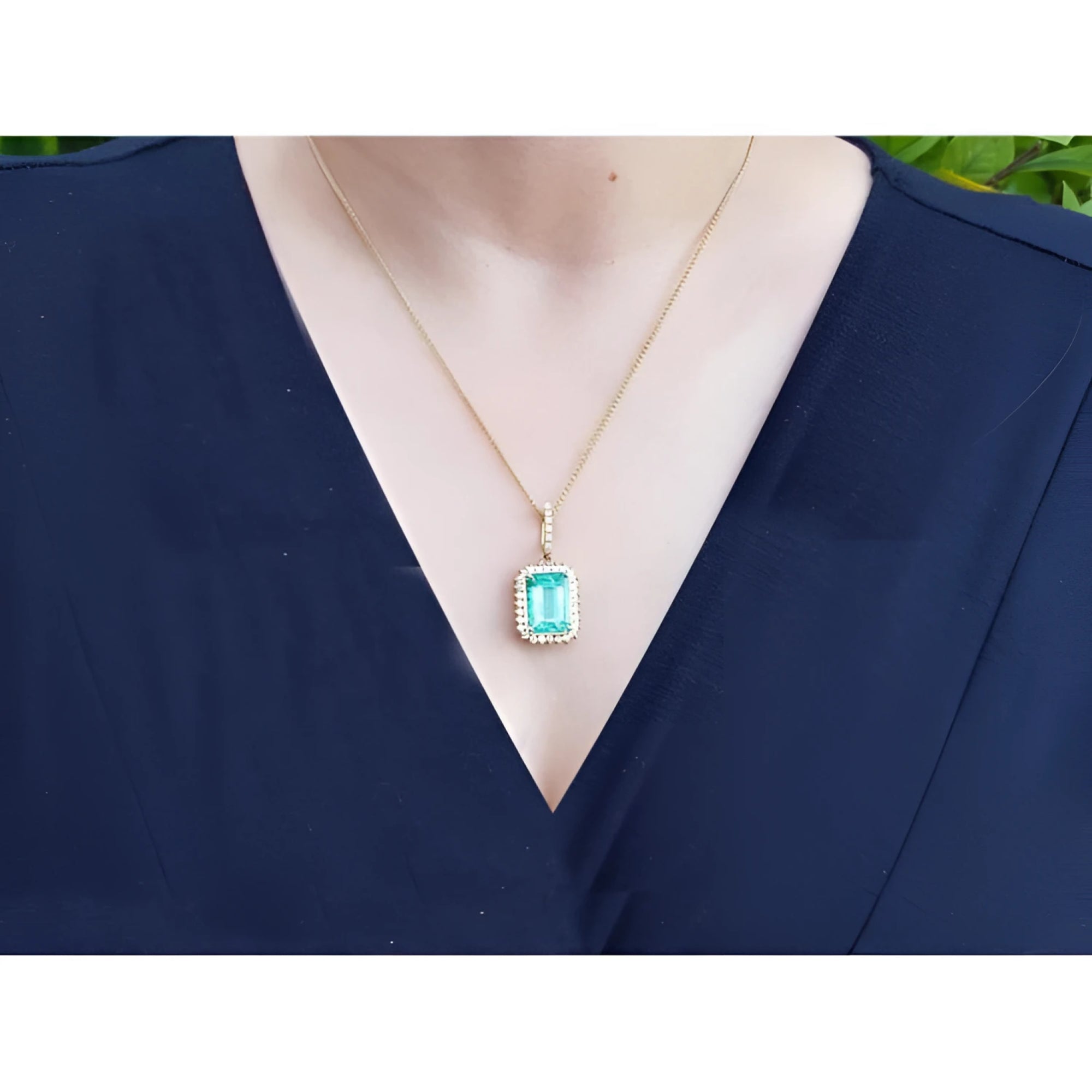 Emerald-cut emerald pendant with halo diamonds on an 18K gold chain, worn on a dark blue top.