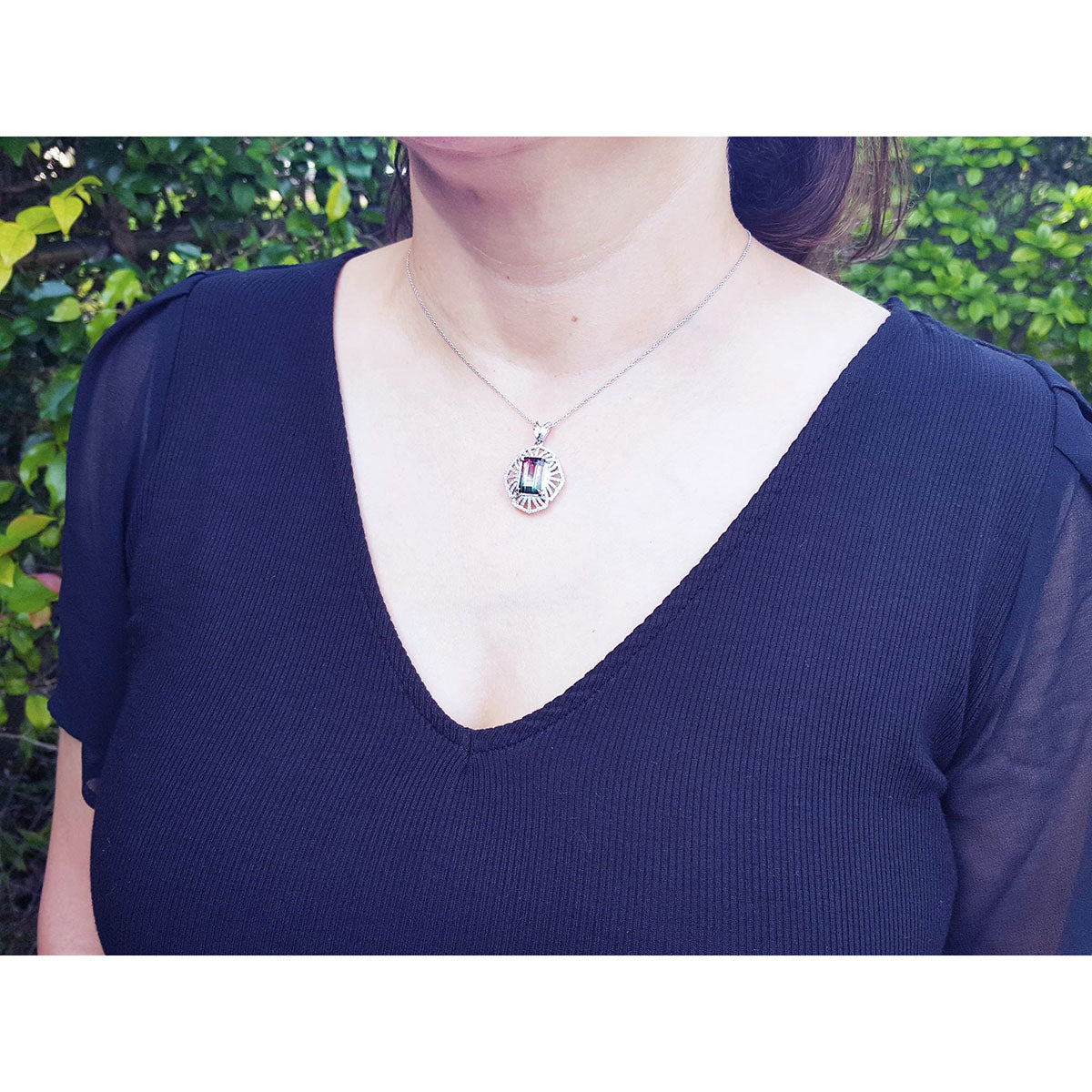 A woman wearing a black top and a white gold chain with a tourmaline pendant on a natural background.