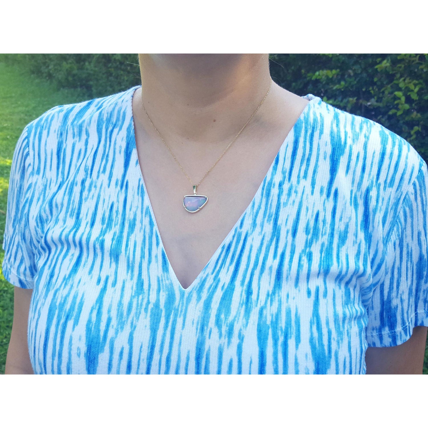 A woman wearing a blue and white patterned top with a gold opal pendant necklace outdoors.